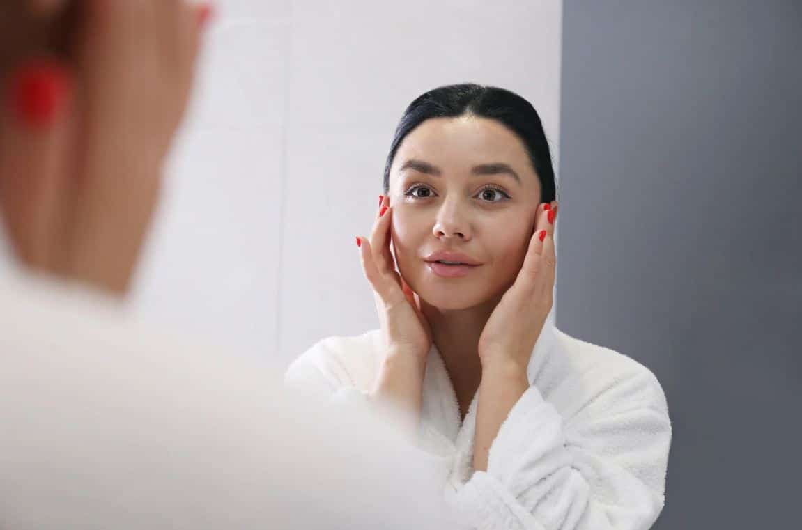 A woman in a white bathrobe is standing in front of a mirror, gently touching her face with both hands. She has long, dark hair and is looking at her reflection with a focused expression. The background features a neutral-colored wall, enhancing the serene atmosphere of her skincare routine. This image conveys themes of self-care, beauty, and personal grooming.