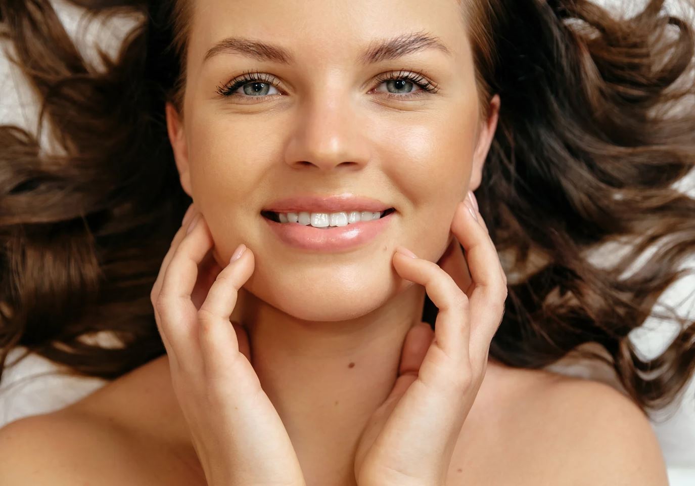 Smiling young woman with long, wavy hair lying on a light-colored surface, gently touching her face with both hands. She has clear skin and bright blue eyes, radiating a sense of happiness and confidence. This image conveys themes of beauty, skincare, and wellness, ideal for promoting beauty products, skincare routines, or self-care practices. The warm lighting enhances her natural features, making it suitable for lifestyle blogs or beauty brands.