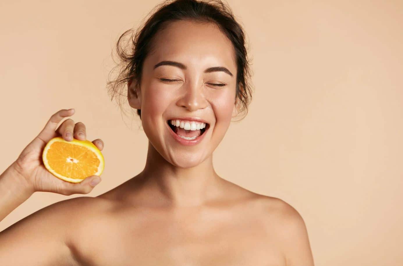 A joyful woman with a radiant smile holds a slice of orange against a soft beige background. Her eyes are closed, and she appears to be laughing, conveying a sense of happiness and vitality. This image emphasizes the refreshing and uplifting qualities of citrus fruits, promoting healthy living and wellness. The natural lighting highlights her glowing skin, making it an ideal representation for topics related to beauty, nutrition, and positivity.