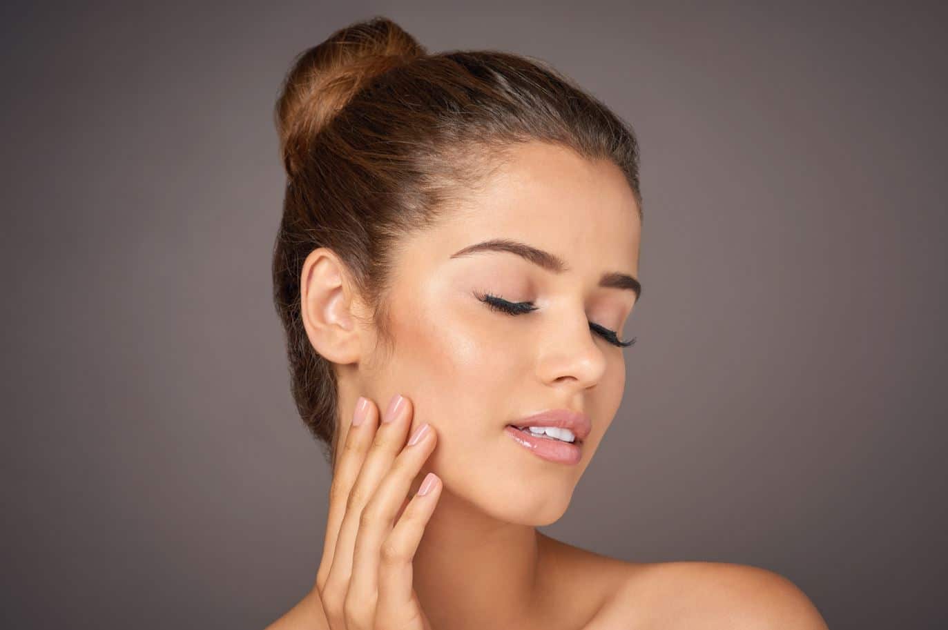 A young woman with a smooth complexion gently touches her face with one hand, showcasing a natural makeup look. Her hair is styled in a neat bun, and she has her eyes closed, exuding a sense of tranquility and confidence. The soft, neutral background complements her glowing skin and enhances the overall aesthetic of beauty and self-care. This image represents skincare, beauty routines, and the importance of self-love and pampering. Ideal for content related to skincare tips, beauty products, or wellness practices.