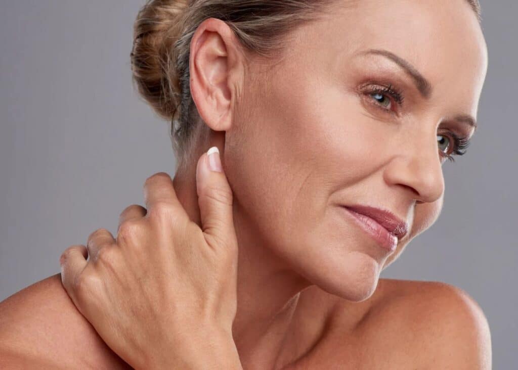 A close-up image of a mature woman gently touching her neck and jawline, showcasing her smooth skin and natural beauty. She has a soft smile and is looking slightly to the side, highlighting her facial features and elegant hairstyle. The background is a subtle gray, enhancing the focus on her expression and skin texture. This image conveys themes of skincare, self-care, and confidence, making it ideal for beauty and wellness content.