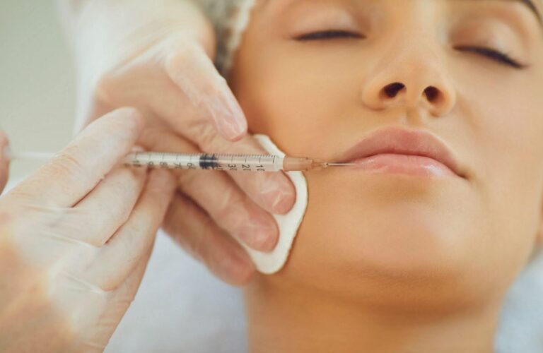 Close-up of a cosmetologist administering a lip filler injection to a patient. The professional, wearing gloves, carefully holds a syringe near the patient&apos;s lips, which are prepped with a cotton pad. The patient has their eyes closed, indicating a relaxed state during the cosmetic procedure. This image highlights the precision and care involved in aesthetic treatments, focusing on enhancing natural beauty through lip augmentation. Ideal for articles on cosmetic procedures, beauty treatments, or skincare.