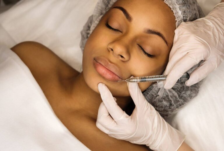 A professional administering a cosmetic lip injection to a relaxed patient in a clinical setting. The patient, with closed eyes, is lying on a treatment table, covered with a white sheet. The practitioner, wearing gloves and a hairnet, carefully holds a syringe near the patient’s lips. This image highlights the process of cosmetic enhancement through injectable treatments, emphasizing safety and professionalism in aesthetic procedures.