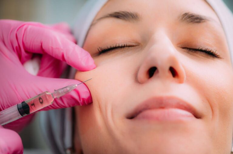 A close-up image of a woman receiving a cosmetic injection in her cheek. The practitioner, wearing pink gloves, is carefully administering the injection with a syringe. The woman appears relaxed with her eyes closed, showcasing a serene expression. This image highlights the process of aesthetic treatments, such as dermal fillers or Botox, aimed at enhancing facial features and reducing wrinkles. It emphasizes the importance of professional care in cosmetic procedures.