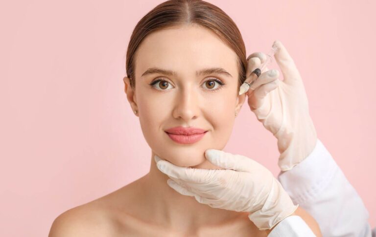 A healthcare professional administers a cosmetic treatment to a woman in a clinical setting. The woman, with a neutral expression, is seated against a soft pink background, highlighting her natural beauty. The professional, wearing gloves, is holding a syringe near her cheek, indicating a procedure such as dermal fillers or Botox. This image represents the growing trend of cosmetic enhancements aimed at achieving youthful and rejuvenated appearances. The focus on safe practices and patient care is evident in the professional&apos;s demeanor and attire.