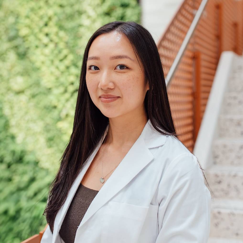 A young woman wearing a white lab coat stands confidently in front of a lush green wall. She has long, straight black hair and a subtle smile, with a delicate necklace visible around her neck. The background features a modern staircase that complements the natural greenery, creating a serene and professional atmosphere. This image conveys a sense of professionalism and approachability, suitable for healthcare or academic contexts.
