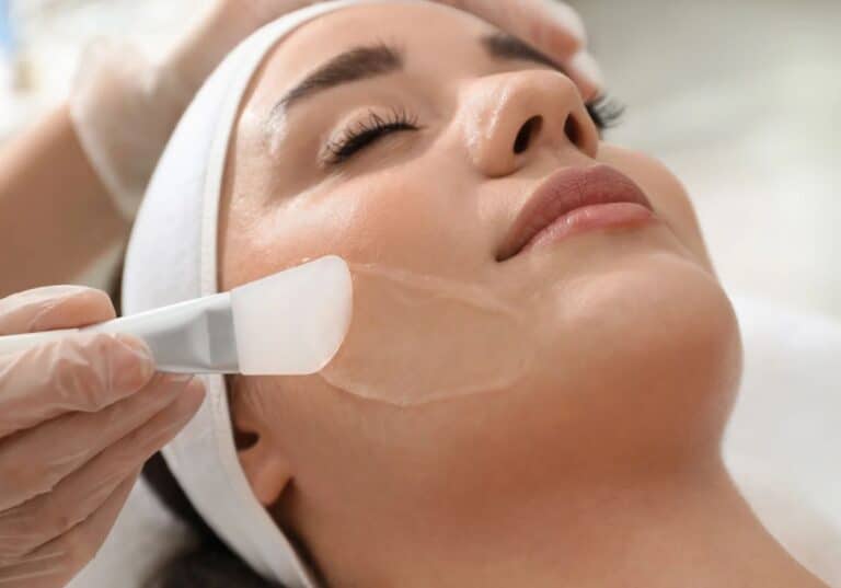 A woman receiving a facial treatment lies comfortably on a treatment bed. A licensed aesthetician, wearing gloves, applies a clear gel mask to her face using a spatula. The woman has a white headband on, and her eyes are closed, indicating relaxation. The setting is a modern skincare clinic with soft lighting, emphasizing a tranquil atmosphere for skincare treatments. This image illustrates the process of facial rejuvenation and highlights the importance of professional skincare for achieving healthy skin.