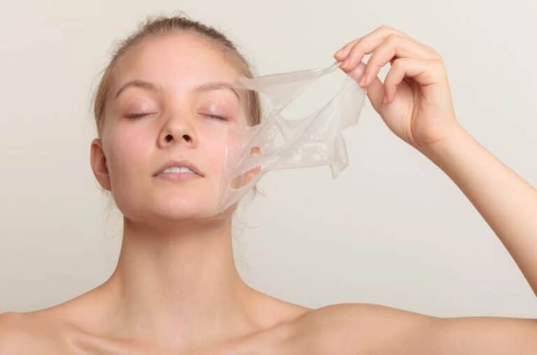 A woman with a serene expression is gently peeling off a transparent facial mask from her face, showcasing a skincare routine. Her eyes are closed, and she appears relaxed as she holds the mask with one hand. The background is a soft, neutral color that highlights her natural beauty. This image emphasizes the importance of self-care and the benefits of using facial masks for healthy skin.