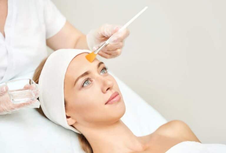 A woman receiving a facial treatment in a spa setting. She is lying on a treatment bed with a white headband and a serene expression. A licensed aesthetician, wearing gloves, is applying a facial product with a brush to her forehead. A small glass container with liquid is visible in the aesthetician&apos;s other hand. The background is neutral and calming, emphasizing relaxation and skincare. This image illustrates a professional skincare procedure focused on rejuvenation and wellness.