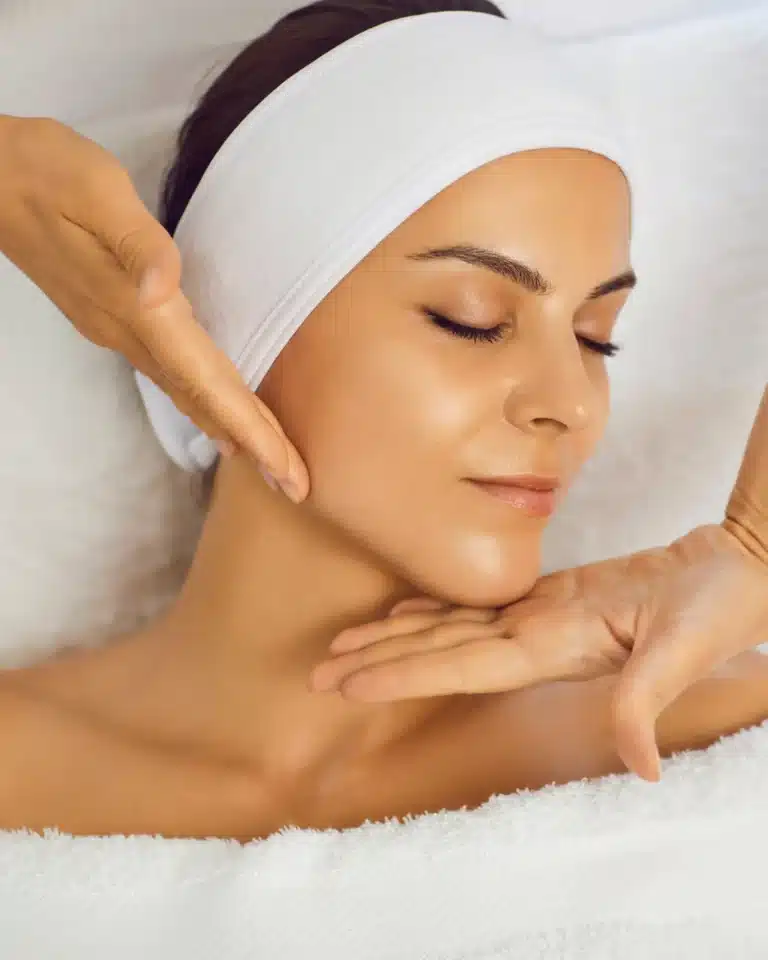 A woman receiving a facial treatment in a spa setting, with her eyes closed and a serene expression. She has a white headband on her hair and is resting on a soft towel. A therapist&apos;s hands gently support her chin, indicating a relaxing skincare routine. The image conveys a sense of tranquility and self-care, highlighting the benefits of professional skincare treatments for healthy, radiant skin.
