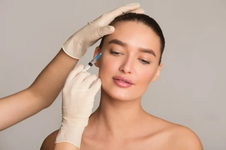 A healthcare professional wearing gloves administers a cosmetic injection to a young woman. The woman appears relaxed, with a neutral expression, as the injector focuses on the area near her eyebrow. The background is a soft gray, emphasizing the procedure&apos;s clinical nature. This image illustrates the process of aesthetic treatments, such as Botox or dermal fillers, highlighting the importance of professional care in cosmetic procedures.