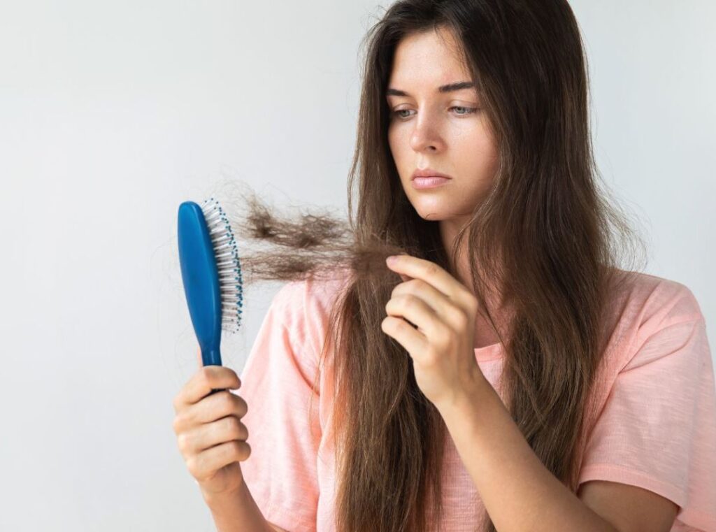 A young woman with long, dark hair examines a tangled section while holding a blue hairbrush in her right hand. She appears focused and contemplative, wearing a light pink shirt. The image captures the struggle of detangling hair, highlighting common hair care challenges. The neutral background emphasizes her expression and the hairbrush, making it relatable for those seeking hair care tips or solutions for managing unruly hair. This visual can be useful for articles or guides on hair maintenance, styling, or product recommendations.