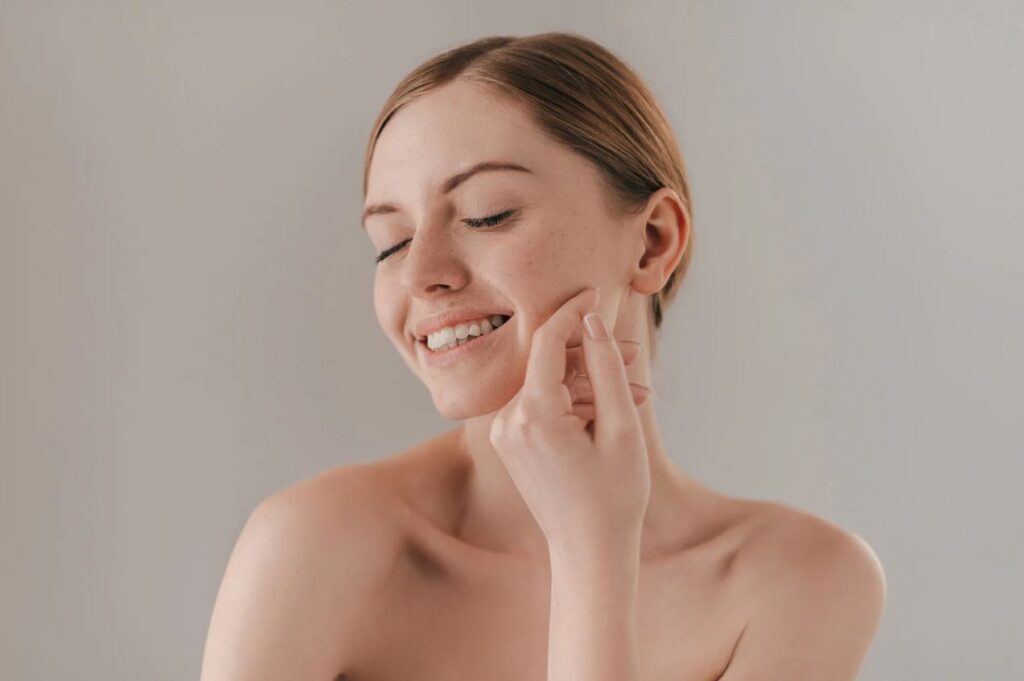 A young woman with light brown hair smiles gently while touching her cheek, showcasing clear skin with a few freckles. She is in a relaxed pose against a soft gray background, highlighting a natural and joyful expression. The image emphasizes skincare and beauty, making it ideal for topics related to skincare routines, self-care, and wellness.