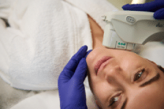 A woman receiving a skincare treatment while lying on a treatment table. She is wearing a white robe and has her hair pulled back. A professional in purple gloves is using a handheld device on her chin, indicating a facial procedure. The setting is a calm and clinical environment, designed for skincare treatments, emphasizing health and wellness.