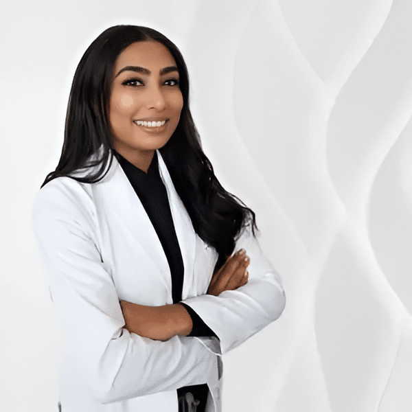 A smiling professional woman wearing a white lab coat stands confidently with her arms crossed. She has long, dark hair and is posed against a soft, textured white background, suggesting a clean and modern environment. This image conveys professionalism and approachability, suitable for a healthcare or wellness setting.