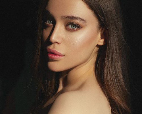 Close-up portrait of a woman with long, straight brown hair and striking blue eyes, featuring natural makeup that highlights her cheekbones and lips. The image captures soft lighting that accentuates her features against a dark background. Ideal for beauty and fashion content, this photo emphasizes elegance and style.